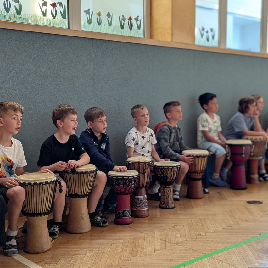 Mehrere Jungen sitzen in einer Reihe, jeder hält eine Trommel. Sie scheinen sich in einem Klassenzimmer mit Holzboden und grauen Wänden zu befinden. Einige haben Blumen am Fenster.