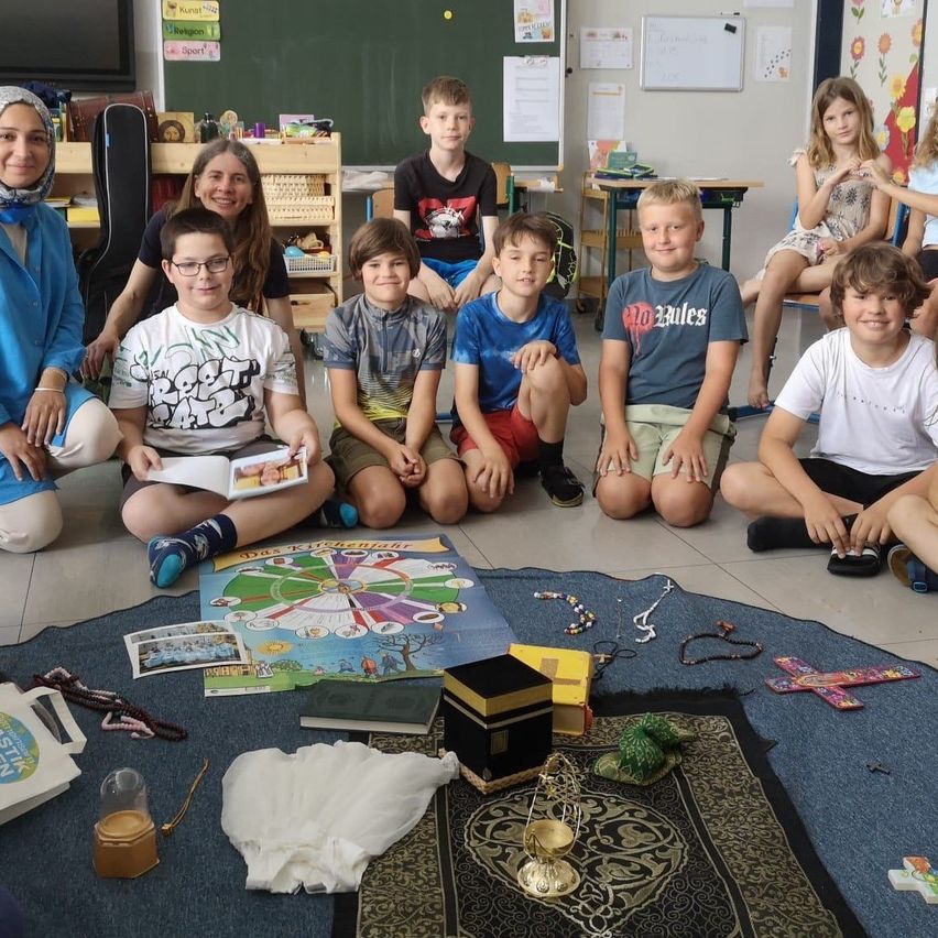 Ein Klassenzimmer mit Kindern auf dem Boden sitzend, einige mit Büchern und einem Brettspiel. Eine Lehrerin lächelt und sitzt neben ihnen.