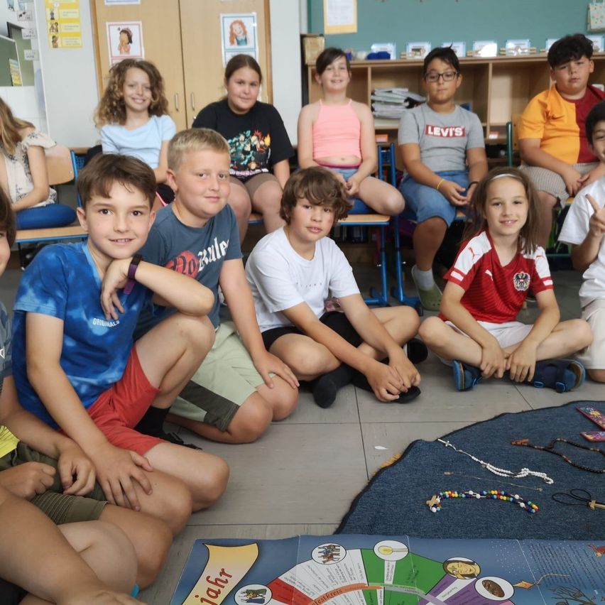 Eine Gruppe von Kindern sitzt im Klassenzimmer auf dem Boden. Sie lächeln und posieren für ein Foto. Einige tragen Armbänder.