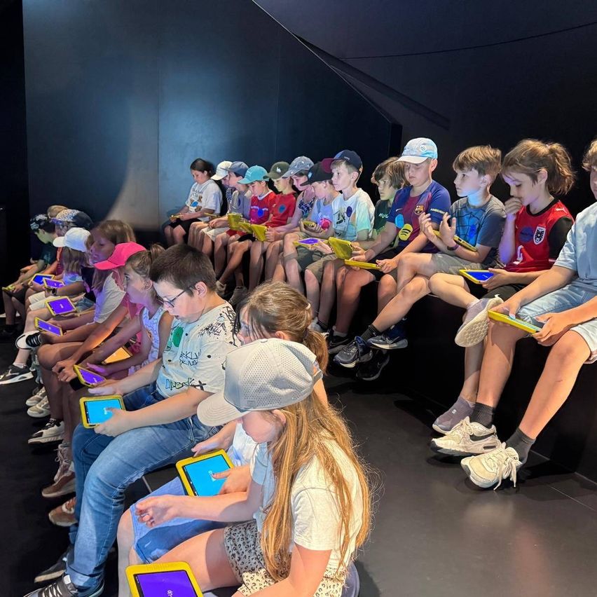 Eine Gruppe von Kindern sitzt in einem dunklen Raum, jeder hält ein Tablet. Sie scheinen engagiert zu sein, einige tragen Mützen und Turnschuhe.