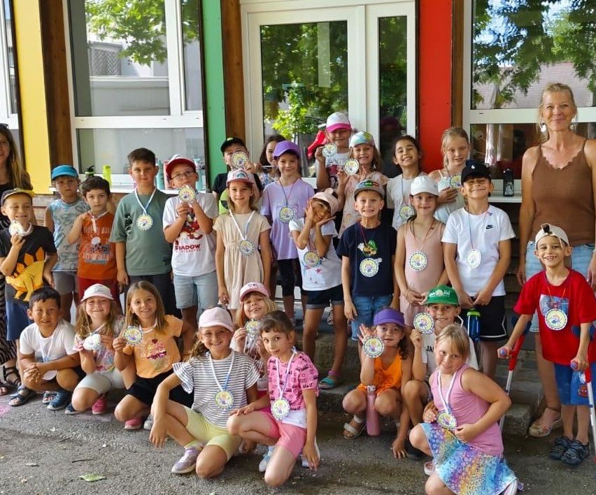 Eine Gruppe von Kindern mit Medaillen um den Hals und einer Frau in einem braunen Tanktop steht vor einem Gebäude.