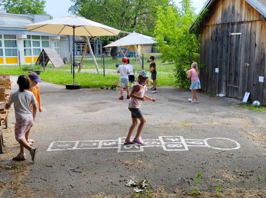 Kinder spielen Hüpfspiel auf einer Straße in einem Spielplatz mit einem Gebäude, einem Zaun und Bäumen im Hintergrund.