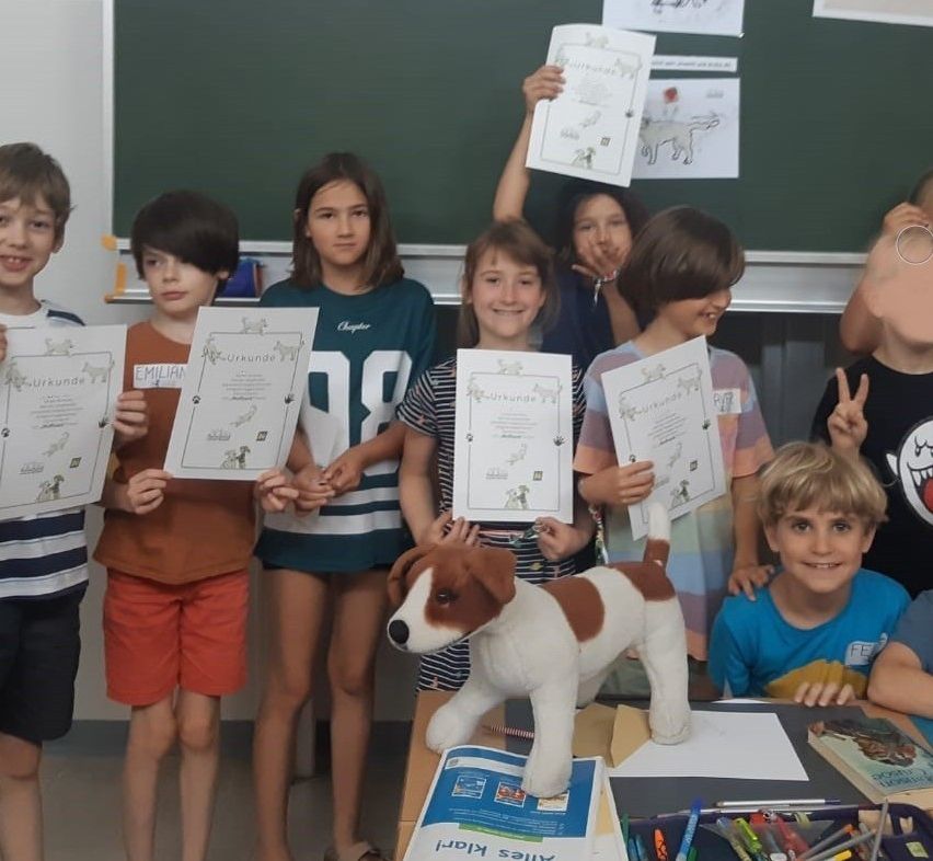Mehrere Kinder stehen in einem Klassenzimmer, halten Urkunden und lächeln. Ein Junge gibt ein Friedenszeichen. Ein Plüschhund sitzt auf einem Schreibtisch.