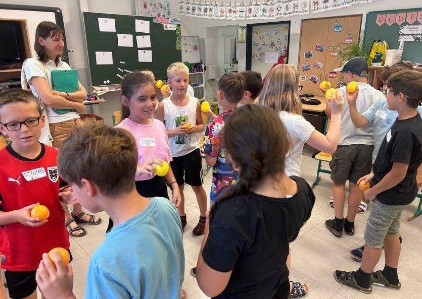In einem Klassenzimmer halten Kinder gelbe Bälle, während eine Frau mit einer grünen Mappe hinter ihnen steht. Tafeln und Pflanzen schmücken die Wände.
