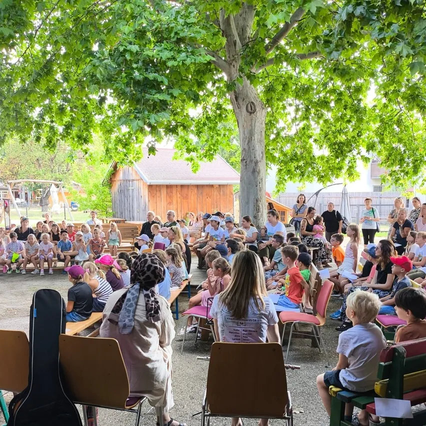Eine Gruppe von Kindern und Erwachsenen ist unter einem großen Baum versammelt und hört einer Präsentation zu. Einige sitzen auf Bänken, andere stehen. Eine Frau vorne hält ein Stück Papier. Hinter ihnen befinden sich Gebäude und ein Spielplatz.