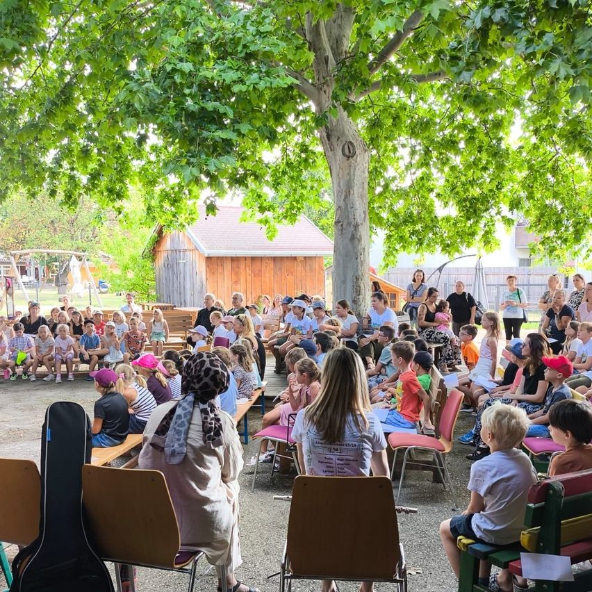 Eine Gruppe von Kindern und Erwachsenen ist unter einem großen Baum versammelt und hört einer Präsentation zu. Einige sitzen auf Bänken, andere stehen. Eine Frau vorne hält ein Stück Papier. Hinter ihnen befinden sich Gebäude und ein Spielplatz.