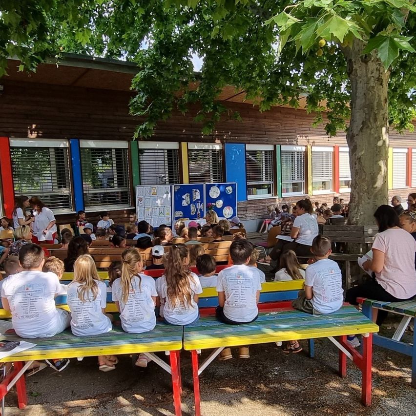 Kinder in weißen T-Shirts sitzen auf Bänken vor einem Gebäude mit farbenfrohen Fenstern. Ein Baum spendet Schatten.