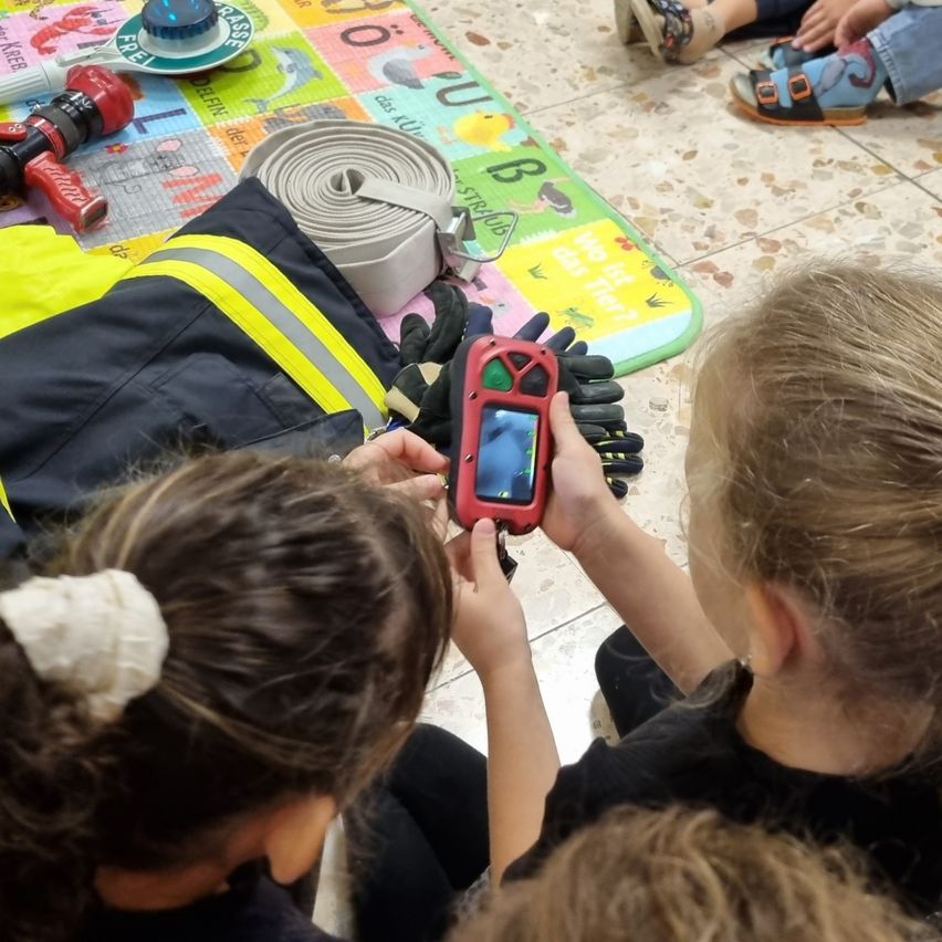 Zwei Kinder untersuchen Feuerwehrausrüstung, einschließlich eines roten Handgeräts, während sie auf dem Boden sitzen.