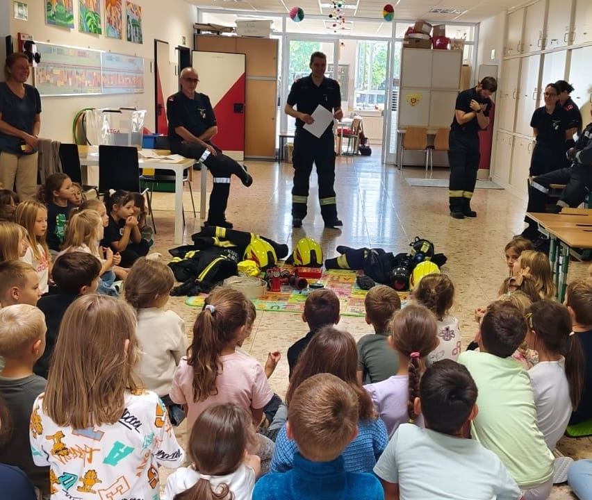 Eine Gruppe von Kindern sitzt im Kreis und hört einem Mann in Feuerwehruniform zu.
