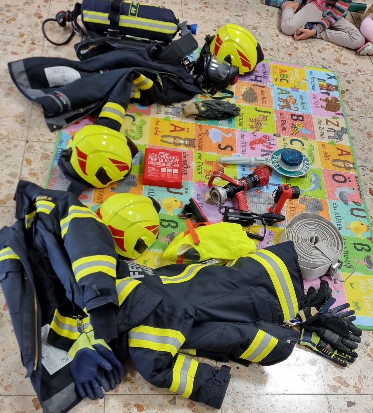 Feuerwehrschutzkleidung, Helme und Ausrüstung liegen auf einer Spielmatte mit Tier-Motiven.
