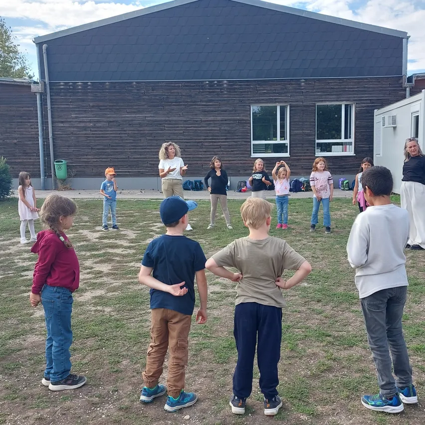 Eine Gruppe von Kindern steht in einem Kreis auf einem Rasen vor einem Gebäude.