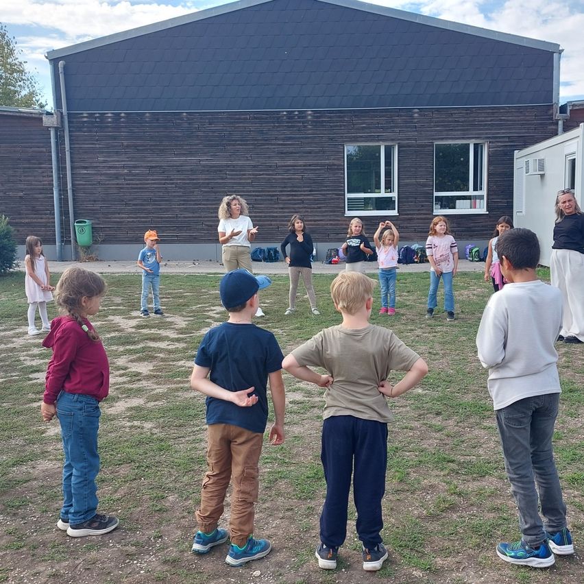 Eine Gruppe von Kindern steht in einem Kreis auf einem Rasen vor einem Gebäude.