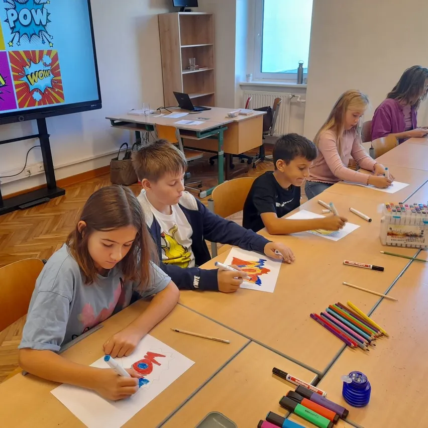 Eine Gruppe von Kindern sitzt an einem Tisch im Klassenzimmer und zeichnet mit Markern auf Papier.
