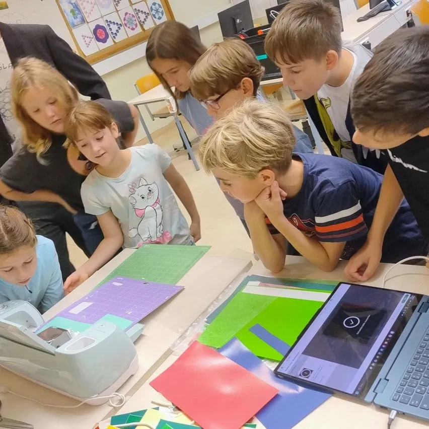 Eine Gruppe von Kindern und Erwachsenen versammelt sich um einen Tisch in einem Klassenzimmer. Sie schauen auf einen Laptop und einen Drucker auf dem Tisch.