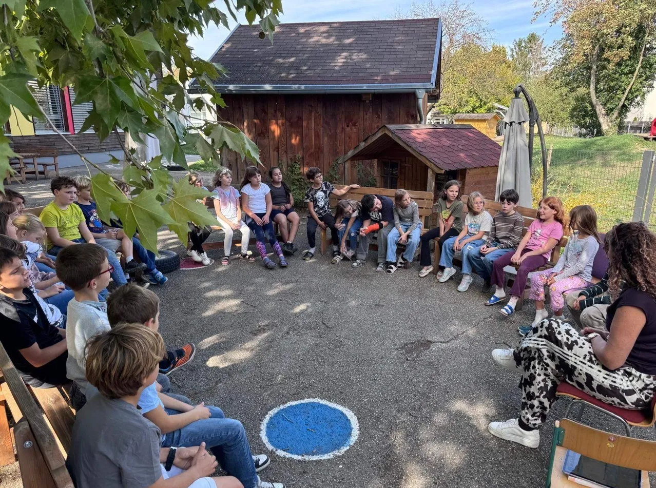 Eine Gruppe von Kindern sitzt im Kreis um einen blauen Kreis auf dem Boden, wahrscheinlich in einem Schulhof.