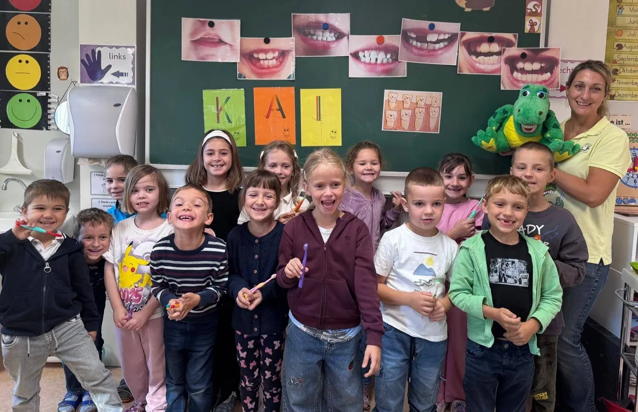 Eine Gruppe von Kindern im Klassenzimmer, alle mit Zahnbürsten in der Hand und lächelnd. Eine Lehrerin steht hinter ihnen mit einem Dinosaurier-Spielzeug.