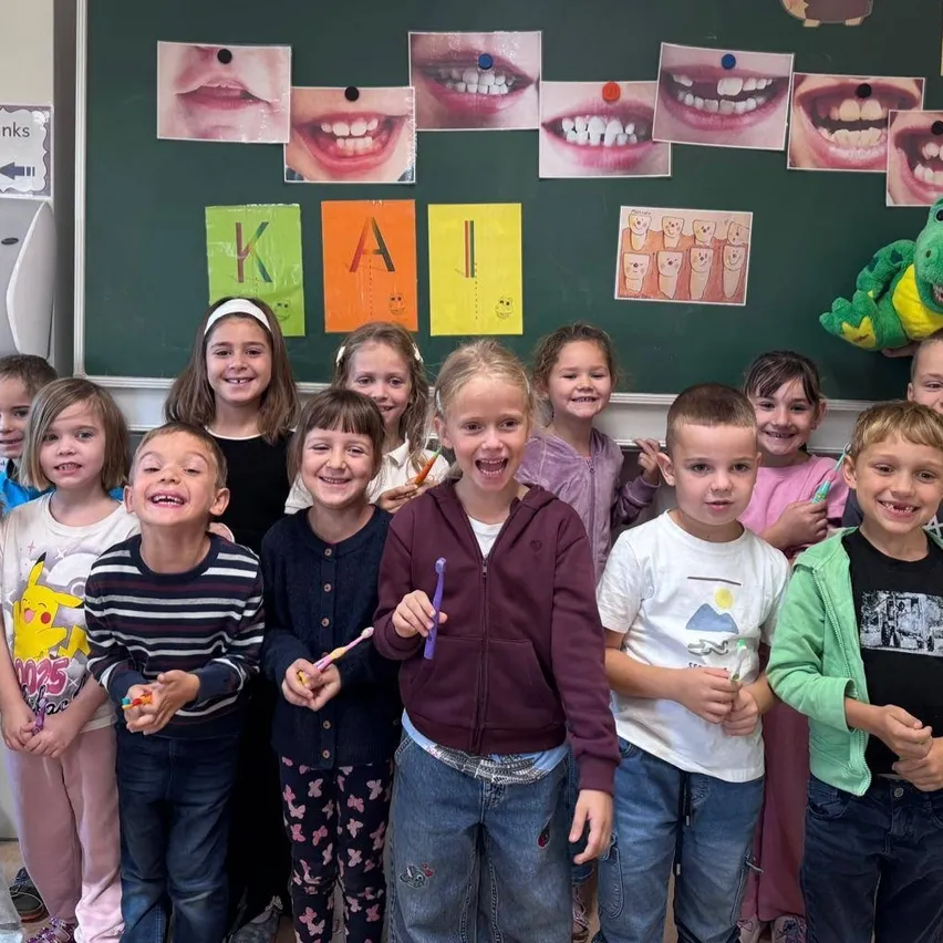 Eine Gruppe von Kindern im Klassenzimmer, alle mit Zahnbürsten in der Hand und lächelnd. Eine Lehrerin steht hinter ihnen mit einem Dinosaurier-Spielzeug.