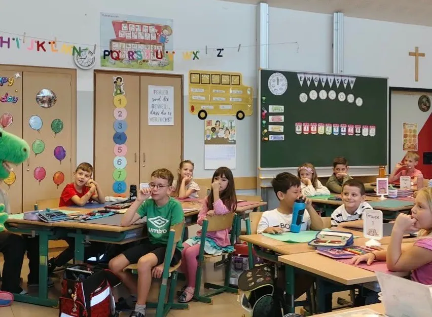 Eine Frau hält ein grünes Dinosaurier-Plüschtier vor einem Klassenzimmer voller Kinder.