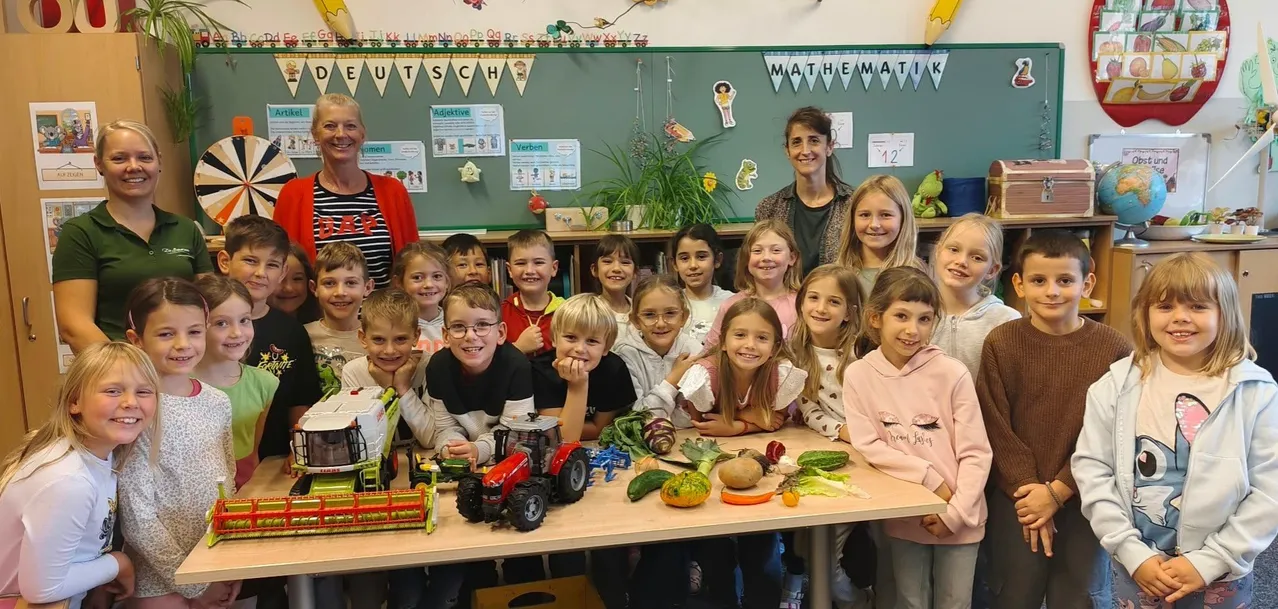 Eine Gruppe von Kindern und zwei erwachsenen Frauen stehen um einen Tisch herum, der mit Gemüse und Spielzeugtraktoren gefüllt ist, in einem Klassenzimmer.