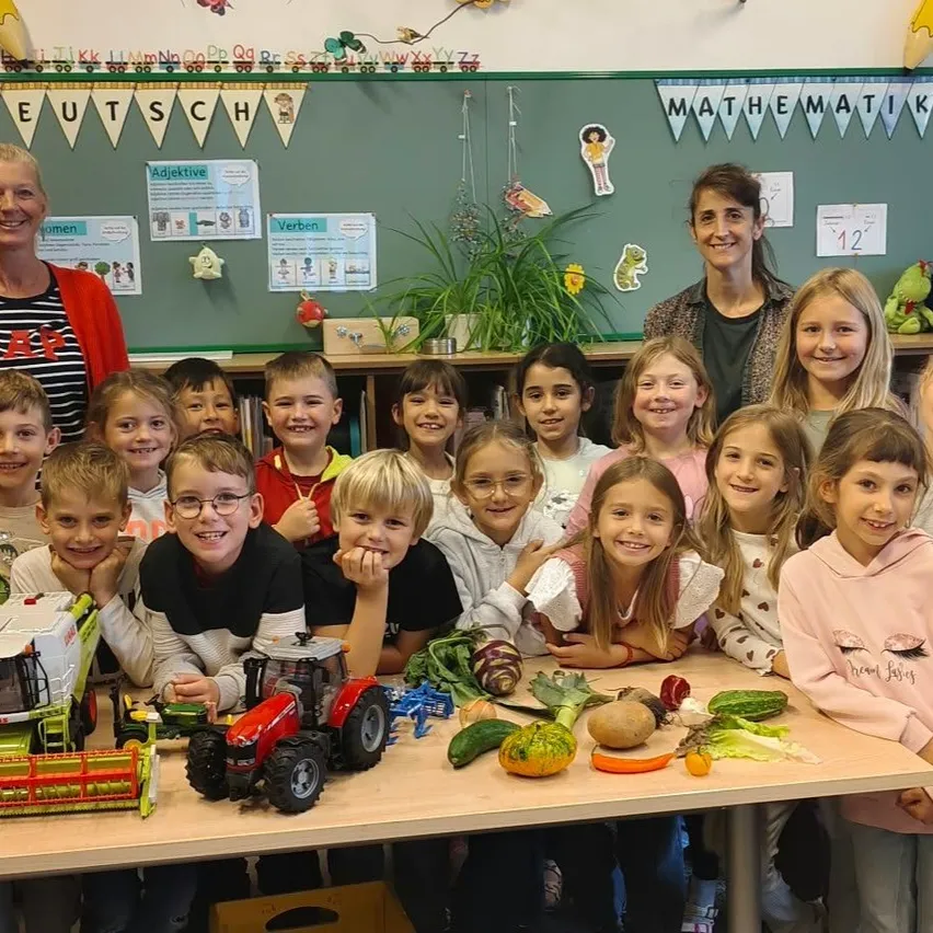 Eine Gruppe von Kindern und zwei erwachsenen Frauen stehen um einen Tisch herum, der mit Gemüse und Spielzeugtraktoren gefüllt ist, in einem Klassenzimmer.
