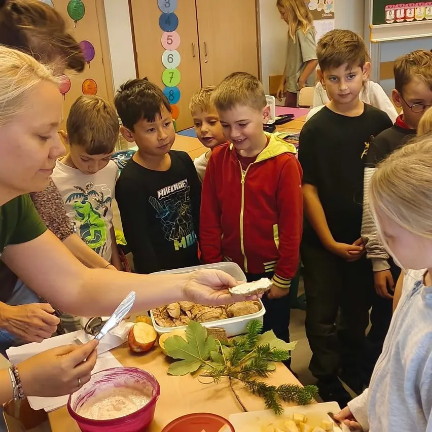 Eine Frau in einem grünen Shirt unterrichtet eine Gruppe von Kindern in einem Klassenzimmer. Sie schneidet Essen auf einem Tisch, während die Kinder zusehen.