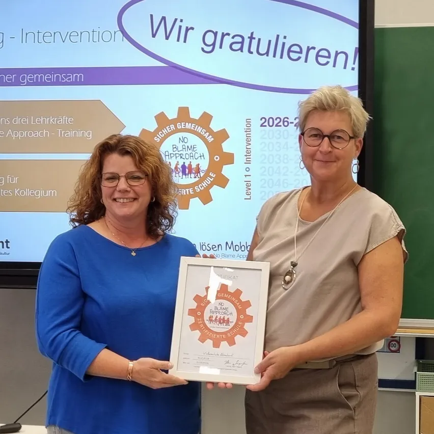 Zwei Frauen stehen vor einem Bildschirm, der eine Präsentation über Mobbing und Intervention zeigt. Die Frau auf der linken Seite hält ein Zertifikat und die Frau auf der rechten Seite lächelt und hält den Rahmen des Zertifikats.