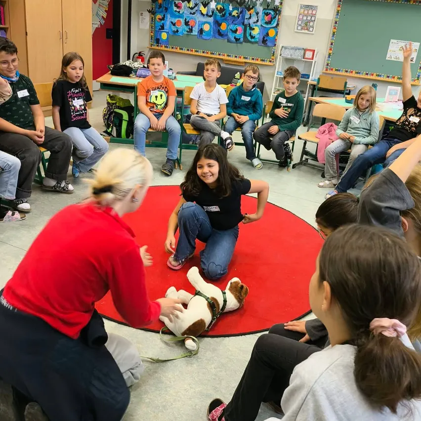 Eine Gruppe von Kindern sitzt im Kreis um eine Frau und einen Hund. Die Frau kniet auf dem Boden und interagiert mit dem Hund, während die Kinder aufmerksam zuschauen.