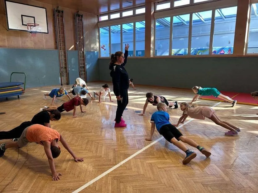 Eine Gruppe von Kindern macht Liegestütze in einer Turnhalle unter der Anleitung einer erwachsenen Frau.