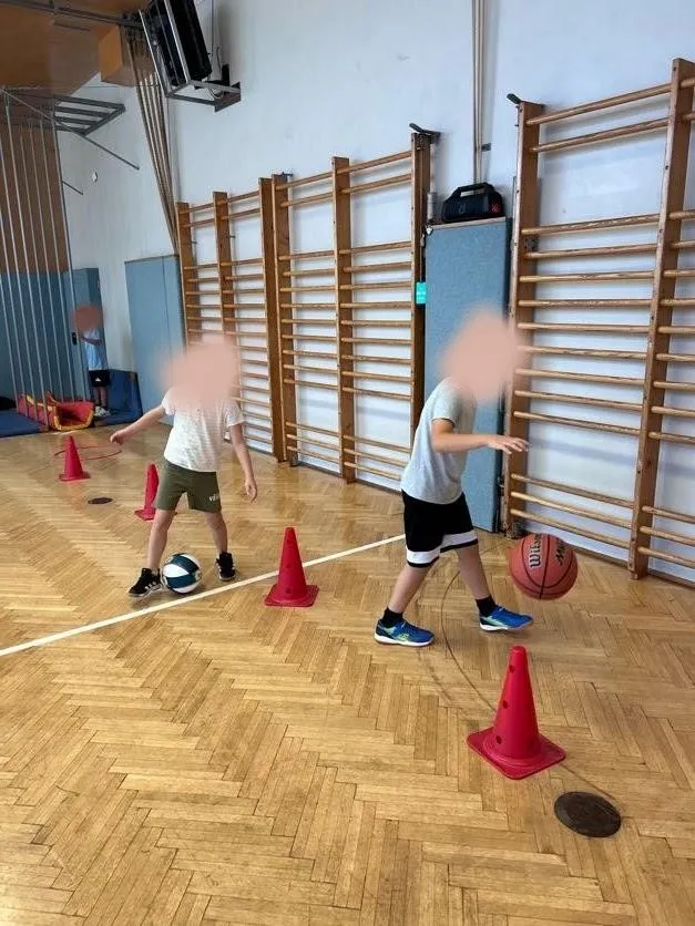 Zwei Kinder spielen Basketball in einer Turnhalle. Ein Kind dribbelt den Ball, während das andere versucht zu blocken.