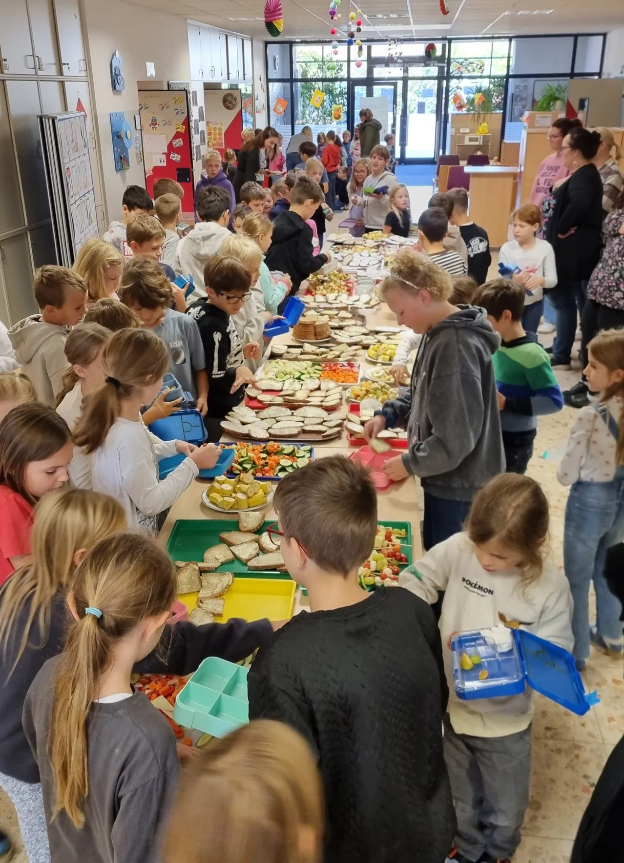 Eine große Gruppe von Kindern versammelt sich um einen langen Tisch, der mit verschiedenen Speisen gefüllt ist. Einige stehen, andere sitzen.