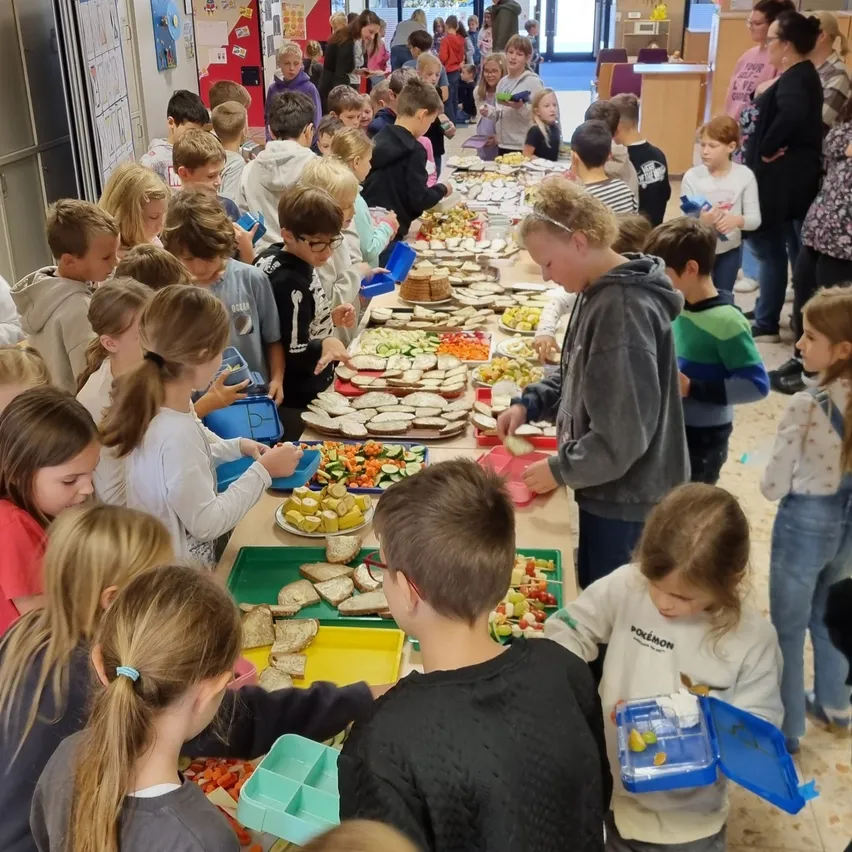 Eine große Gruppe von Kindern versammelt sich um einen langen Tisch, der mit verschiedenen Speisen gefüllt ist. Einige stehen, andere sitzen.