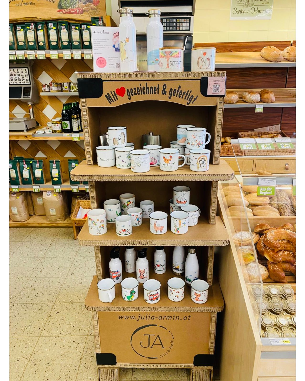 Ein Karton-Display mit verschiedenen Bechern und Flaschen ist in einem Geschäft. Ein Schild sagt 'Mit Liebe gezeichnet & bedruckt.'