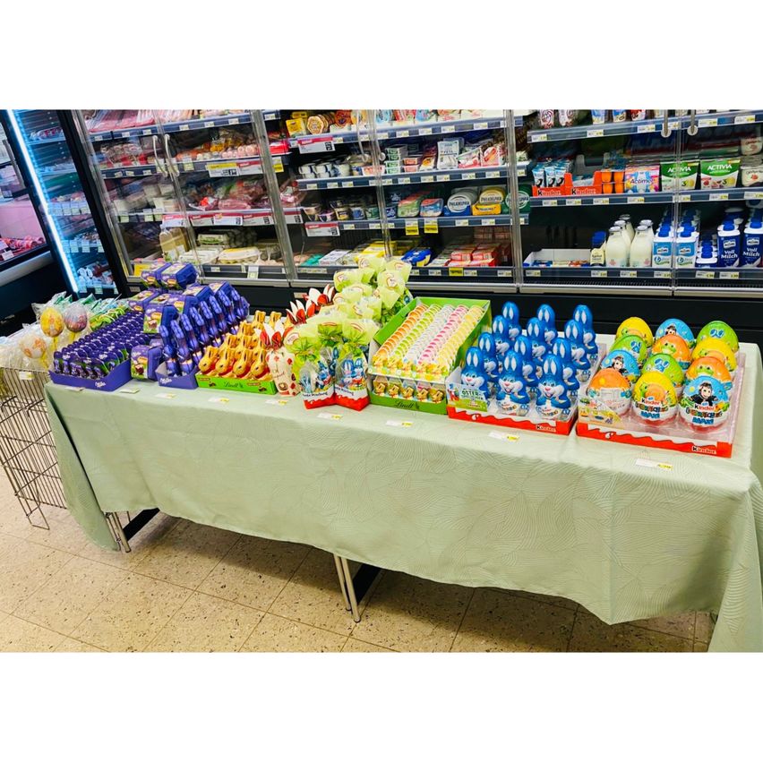 Ein Supermarktregal zeigt eine Vielzahl von Oster-Produkten, einschließlich Schokoladeneiern, Süßigkeiten und Spielzeug, ordentlich auf einem Tisch mit grüner Tischdecke angeordnet.