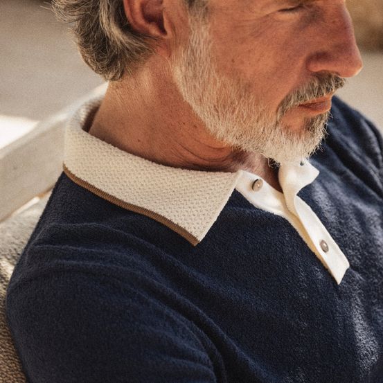 Eine Nahaufnahme eines älteren Mannes in einem dunkelblauen Poloshirt mit weißem Kragen und goldenem Besatz. Er hat einen weißen Bart und Schnurrbart.