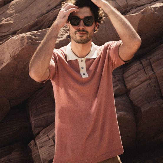 Ein Mann steht im Freien, justiert seine Sonnenbrille und trägt ein rosa Poloshirt und braune Hose vor einem Hintergrund großer, felsiger Ausläufer.