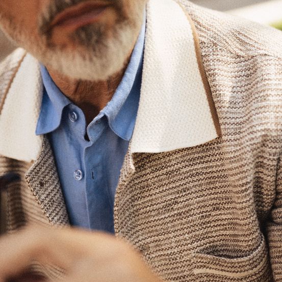 Ein Mann mit weißem Bart und Schnurrbart trägt ein hellblaues Hemd und eine braune gestrickte Jacke mit weißem Besatz.