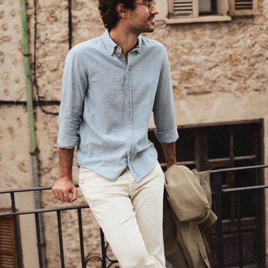 Ein Mann steht auf einem Balkon, trägt ein hellblaues Hemd und weiße Hosen und hält einen beigen Mantel. Hinter ihm steht ein Gebäude aus Stein mit Fenstern.