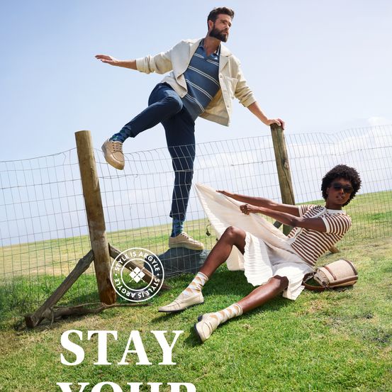 Zwei Personen in lässiger Kleidung, eine steht auf einem Zaun und die andere sitzt auf Gras. Beide tragen Schuhe und sind von einem Zaun und Gras umgeben.