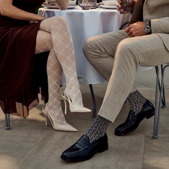 Eine Frau in einem kastanienbraunen Kleid und ein Mann in einem beigen Anzug sitzen zusammen auf einem Stuhl in der Nähe eines Tisches. Der Mann trägt gemusterte Socken und die Frau weiße Absätze.