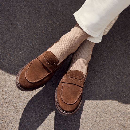 Eine Person, die braune Wildleder-Loafer und beige Socken mit strukturierten Mustern auf einer grauen Oberfläche trägt.