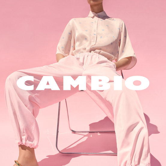 Eine Frau sitzt auf einem Stuhl vor einem rosa Hintergrund. Sie trägt ein rosa Hemd und eine Hose. Das Wort 'CAMBIO' ist auf dem Bild geschrieben.