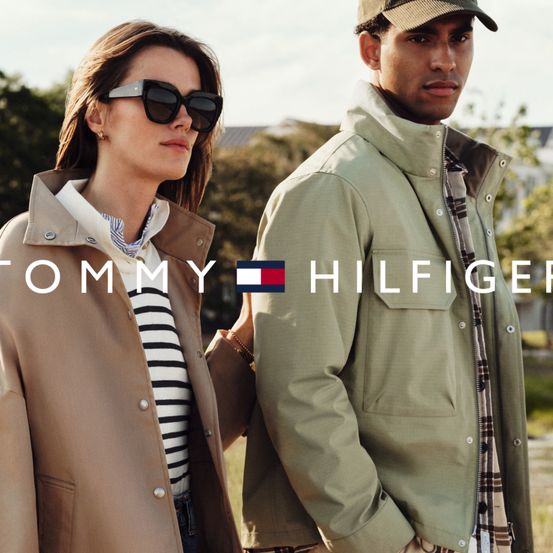 Ein Mann und eine Frau, beide in Tommy Hilfiger-Kleidung, stehen draußen. Die Frau trägt einen beigen Mantel und eine Sonnenbrille, der Mann eine grüne Jacke.