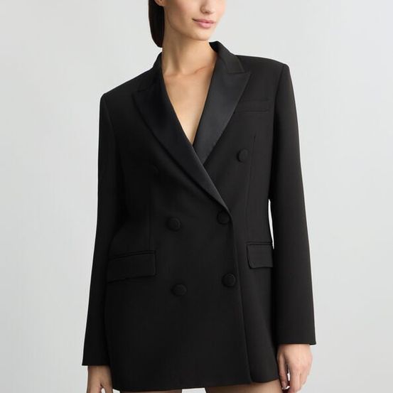 Eine Frau trägt einen schwarzen, doppelreihigen Blazer mit schlanker Passform und steht mit den Händen auf den Hüften.