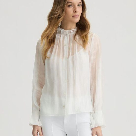 Eine Frau in weißer Kleidung posiert vor einem schlichten Hintergrund. Sie trägt ein weißes Oberteil mit langen Ärmeln und einem Rüschenkragen. Ihr Haar ist lang und offen.