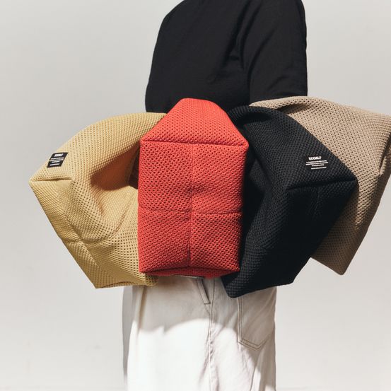 Eine Person in einem schwarzen Shirt hält einen Stapel von Kissen: beige, rot und schwarz. Das rote Kissen hat einen weißen Aufkleber.