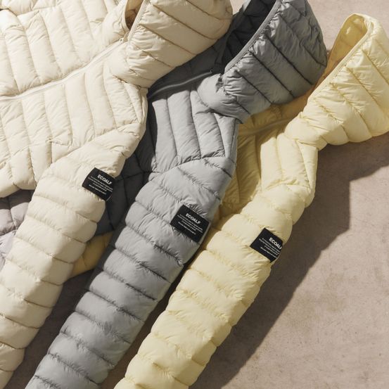 Drei umweltfreundliche Daunenjacken in Weiß, Hellgrau und Gelb sind auf einer strukturierten Oberfläche ausgestellt. Jede Jacke hat eine Kapuze und einen Reißverschluss.