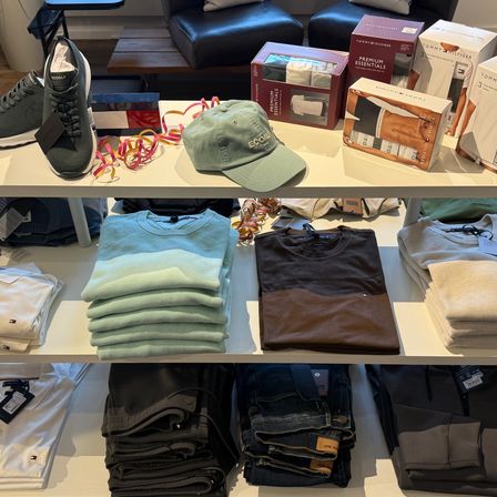 Ein Bekleidungsgeschäft mit einem Ständer gefalteter Hemden, einem braunen T-Shirt, einem Stapel Jeans und einer Box Tommy Hilfiger Essentials auf einem Regal.
