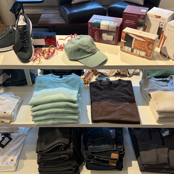 Ein Bekleidungsgeschäft mit einem Ständer gefalteter Hemden, einem braunen T-Shirt, einem Stapel Jeans und einer Box Tommy Hilfiger Essentials auf einem Regal.