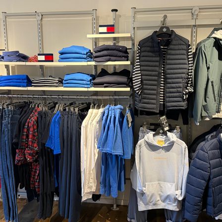 Ein Bekleidungsgeschäft zeigt eine Vielzahl von Kleidungsstücken, darunter Jacken, Hoodies, Hemden und Hosen. Die Jacken hängen an der Wand und die Hoodies befinden sich in den Regalen. Die Kleidung ist ordentlich organisiert und gefaltet. Das Geschäft hat einen modernen und organisierten Look.