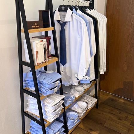 Ein Kleiderständer mit einer Auswahl an Hemden und Krawatten, einige gefaltet, andere hängend, auf Holzregalen an einer weißen Wand.